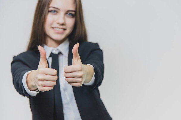 Female hand gestures thumbs up. Business girl is smiling broadly and shares positive emotions. Focus on the thumbs. Background is blurred. - Photo, Image