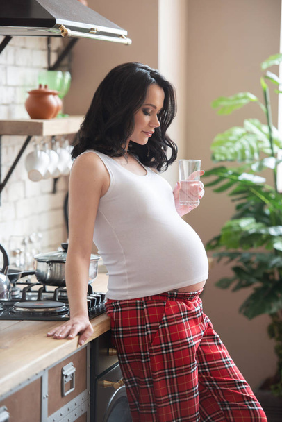 A young beautiful woman in her checkered pajamas is drinking a glass of pure water in the kitchen. Beautiful healthy pregnancy and fresh skin - Foto, Imagen
