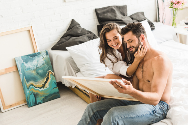 junges Paar sitzt im Bett und lächelt, während Mann Gemälde hält und Mädchen lächeln, während sie Kerl umarmen - Foto, Bild