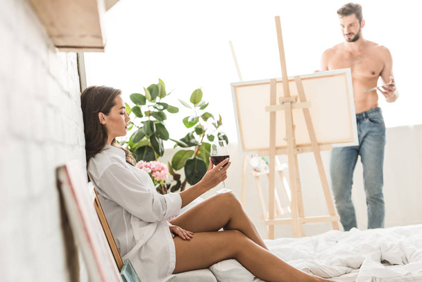 side view of girl sitting in bed with wine while shirtless man drawing girlfriend with brush and colorful paints - Valokuva, kuva