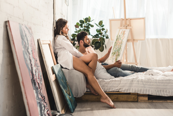 side view of sexy young couple looking at painting while man lying in bed and girl sitting behind guy - Фото, изображение