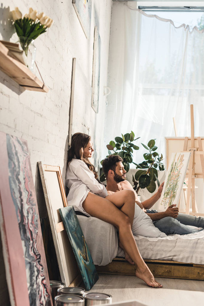 side view of sexy young couple looking at painting while man lying in bed and girl sitting next to guy - Фото, изображение