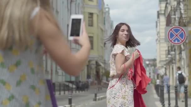 Girl photographs on cell phone happy adorable fun fashionista girlfriend outdoors. Happy girl shows off her new red dress standing on the street together. Leisure of happy young women. - Materiał filmowy, wideo