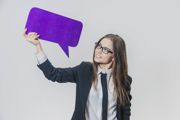 Young woman is holding a violet speech bubble over herself, which is rectangular-shaped, isolated, on the white background. Young lady is looking at speech bubble and is smiling widely. - Foto, Bild