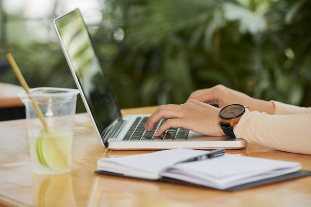 Close-up de mulher sentada à mesa com bloco de notas e limonada fria e digitando no teclado do computador portátil ela trabalha no café
 - Foto, Imagem