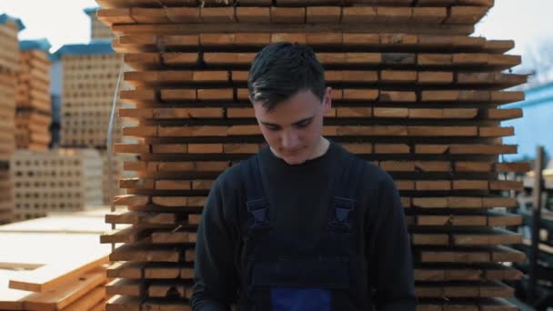 Mladý pracovník nosí stavební googgles a uniformu. Muž se stydlivě dívá na kameru. palet na pozadí. Smečka dřevěných hobulků za ním. - Záběry, video
