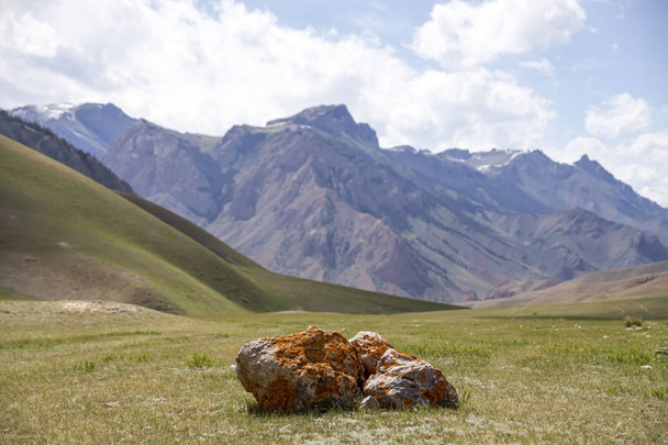Jäkälällä peitetty lohkare laitumella sinistä taivasta vasten pilvillä ja vuorijonoilla. Matkustaa. Kirgisia
 - Valokuva, kuva