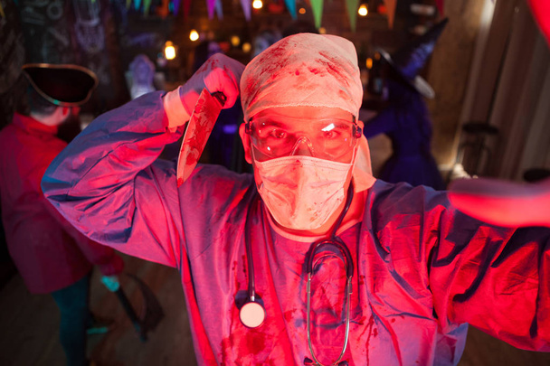 Hombre sosteniendo un cuchillo cubierto de sangre vestido como un médico espeluznante en la fiesta de Halloween
 - Foto, Imagen