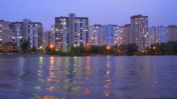 Apartmenthäuser in der Nähe des Sees, bei Sonnenuntergang mit violettem Himmel, der sich im Wasser spiegelt. Abend Stadtbild Hintergrund. Zeitraffer. Abendliche Stadtbeleuchtung, die sich im Wasser, Fluss, See spiegelt. - Filmmaterial, Video