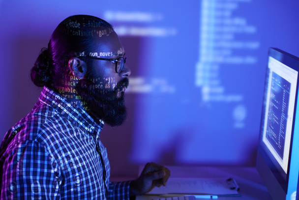 Молодой бородатый программист в очках смотрит на монитор компьютера и изучает программное обеспечение в темном офисе
 - Фото, изображение