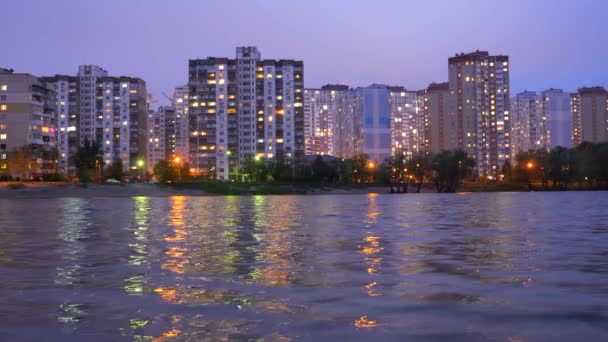 Un complesso di edifici di appartamenti nella città serale vicino al grande lago, con lampioni che si riflettono nell'acqua. Edifici al crepuscolo vicino al lago. Edifici e luci in appartamenti sera
. - Filmati, video