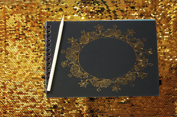 Notizbuch mit leeren schwarzen Blättern und einem hölzernen Schaber auf hellem, glänzendem Hintergrund aus Goldpailletten, Platz für Notizen, Text zur Erinnerung auf schwarzem Papier schreiben - Foto, Bild