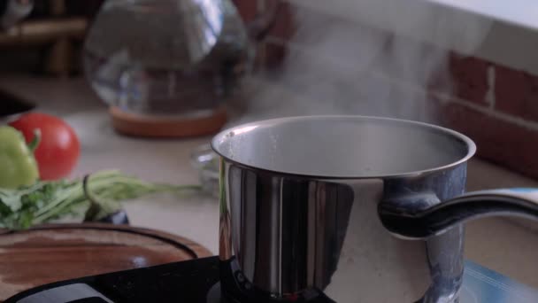 Pan stands on the stove. Boiling water rises steam. Vegetables on a cutting board in the background. - Footage, Video