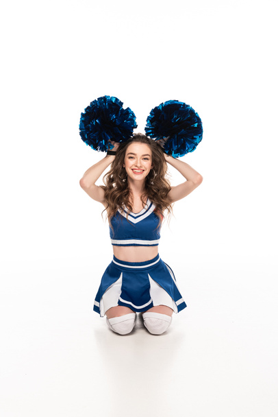 sexy gelukkig cheerleader meisje in blauw uniform zittend op de vloer en het houden van pompoms in de buurt van hoofd geïsoleerd op wit - Foto, afbeelding