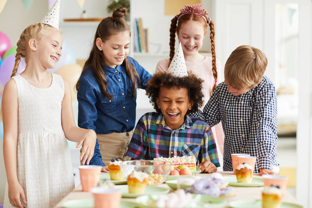 Groupe multi-ethnique d'enfants célébrant anniversaire debout à table avec un garçon afro-américain le tirant visage dans le gâteau
 - Photo, image