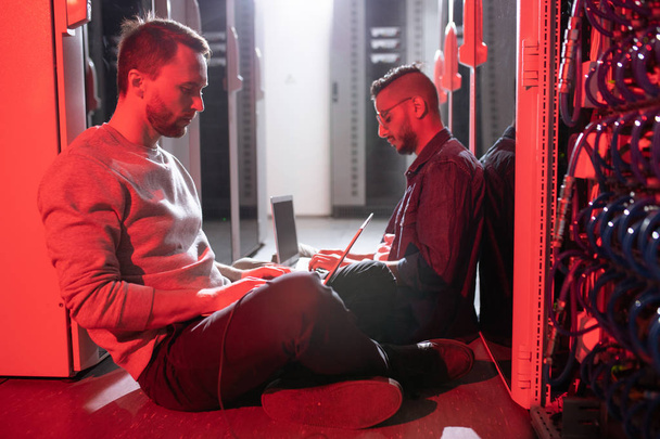 Jóvenes ingenieros multiétnicos concentrados sentados en el suelo en la sala de servidores con luz roja y trabajando con computadoras portátiles
 - Foto, Imagen