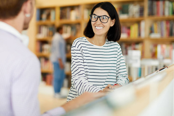 Θετική ενθουσιασμένη νεαρή γυναίκα σε γυαλιά που έχουν τρελό χαμόγελο, ενώ κουβεντιάζοντας με το φίλο στο βιβλιοπωλείο - Φωτογραφία, εικόνα