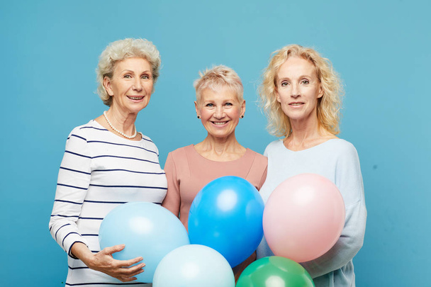 Porträt lächelnder reifer Damen in lässigen Outfits, die zusammen vor blauer Wand stehen und Luftballons in die Höhe halten - Foto, Bild