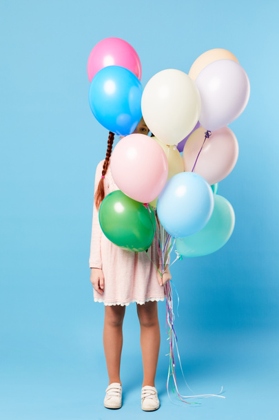 Full length portrait of cute girl hiding behind balloons standing against pastel blue background, party concept, copy space - Photo, Image