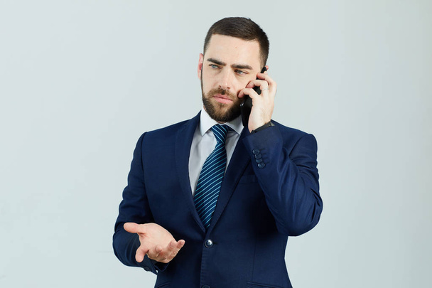 Vakava tyytymätön nuori parrakas liikemies virallisessa puvussa seisoo valkoista seinää vasten ja elehtii käsi puhuessaan matkapuhelimella
 - Valokuva, kuva