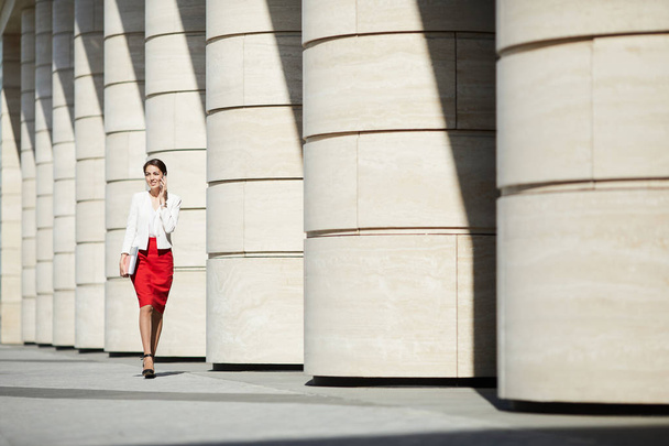 Portrait complet d'une élégante jeune femme portant une jupe rouge marchant le long d'une rangée de piliers et parlant par téléphone, espace de copie
 - Photo, image