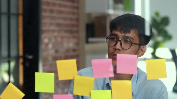 Asian creative man stick a sticky note on glass board. Young professional business male write information, strategy, reminder on paper, business situation, startup in Loft office concept. - Footage, Video