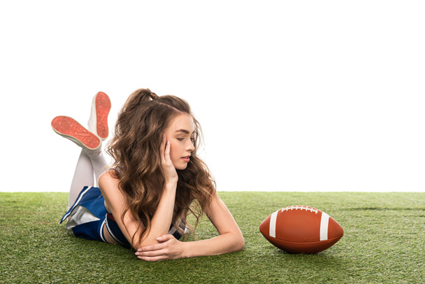 cheerleader girl in blue uniform lying near rugby ball on green field isolated on white - Фото, изображение