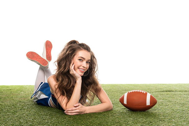 happy cheerleader girl in blue uniform lying near rugby ball on green field isolated on white - Фото, изображение