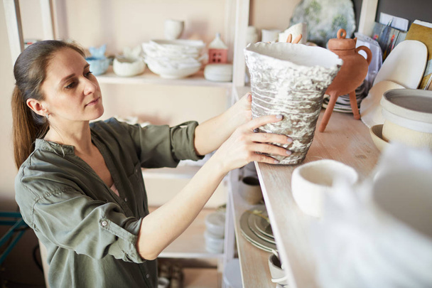 Retrato de vista lateral de alfarera femenina contemporánea poniendo jarrón hecho a mano en el estante en el estudio, espacio de copia
 - Foto, Imagen