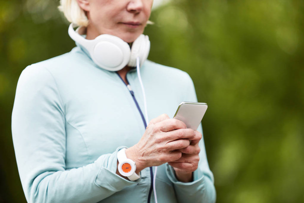Aged sportswoman in activewear texting or scrolling in smartphone for soundtrack in playlist before working out outdoors - Foto, imagen