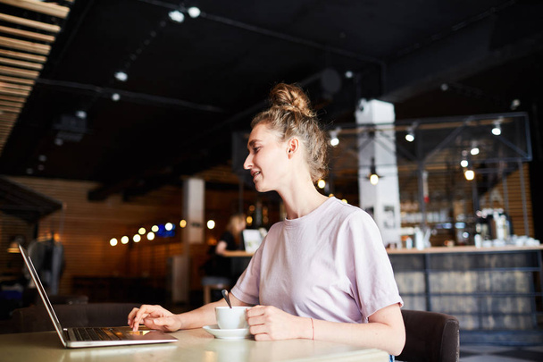 Jovem empreendedora positiva com coque de cabelo sentado à mesa na cafetaria e usando laptop enquanto planeja o projeto de inicialização e bebe café
 - Foto, Imagem