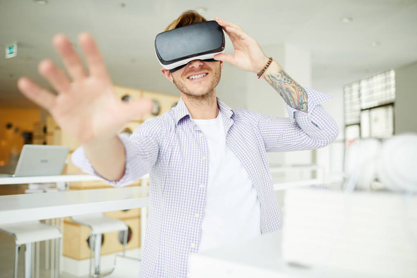 Vrolijke nieuwsgierige hipster kerel met kleurrijke tatoeage ondergedompeld in virtuele wereld die de hand naar de camera reikt terwijl je de VR-headset aanpast - Foto, afbeelding