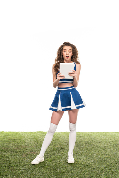 shocked cheerleader girl in blue uniform holding digital tablet on green grass isolated on white - Photo, Image