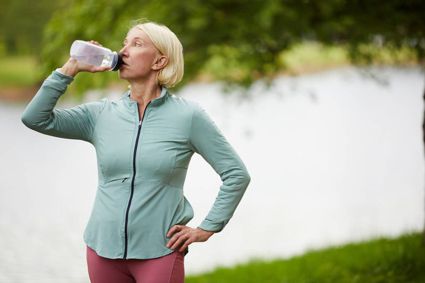 Mature blonde female drinking water from plastic bottle while having rest after workout in natural environment by waterside - Foto, Imagem