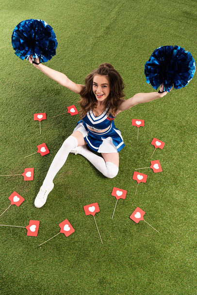 overhead view of happy cheerleader girl in blue uniform sitting with pompoms on green field with scattered likes signs - Foto, Bild