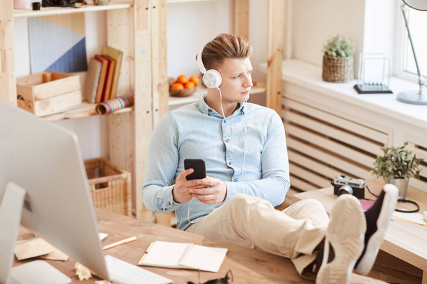 Серйозний пенсійний молодий хлопець в білих навушниках тримає ноги на столі і використовує смартфон, відповідаючи на повідомлення і слухаючи музику через навушники
 - Фото, зображення