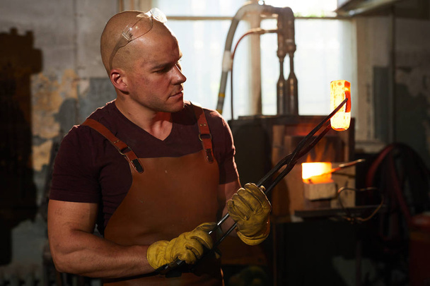 Задумчивый молодой фабричный рабочий в фартуке и перчатках, работающий с обогреваемым металлическим материалом в цехе
 - Фото, изображение