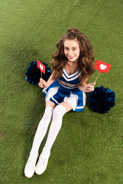 bovenaanzicht van mooie cheerleader meisje in blauw uniform zittend met pompoms en sociale media harten op groen veld - Foto, afbeelding