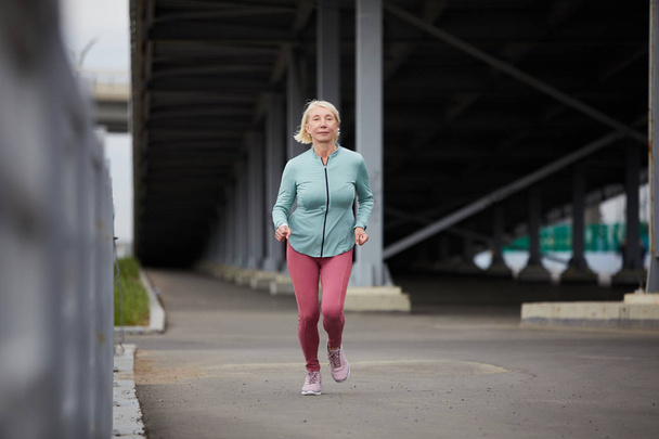 Здоровая блондинка в спортивной одежде бежит по шоссе под мостом во время утренних тренировок
 - Фото, изображение