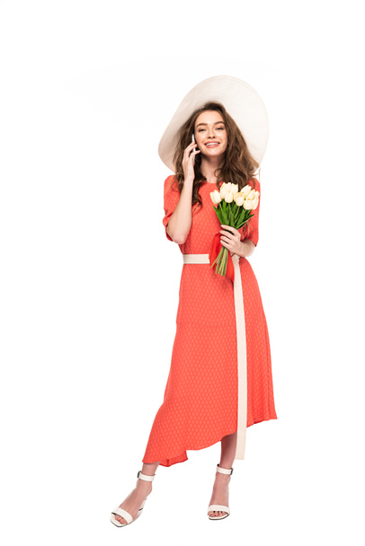 mujer elegante feliz en sombrero y vestido sosteniendo tulipanes blancos y hablando en el teléfono inteligente aislado en blanco
 - Foto, Imagen