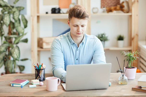 Серьезный занятой молодой человек со стильной прической, сидящий за столом и печатающий на ноутбуке во время работы над отчетом в офисе
 - Фото, изображение