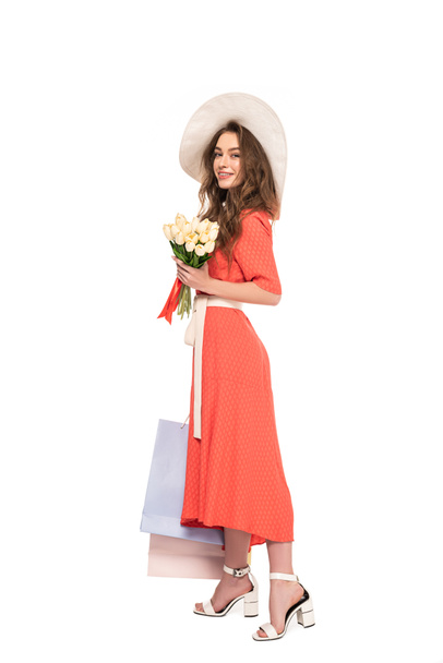 happy elegant woman in hat and dress holding white tulips and shopping bags isolated on white - Foto, afbeelding