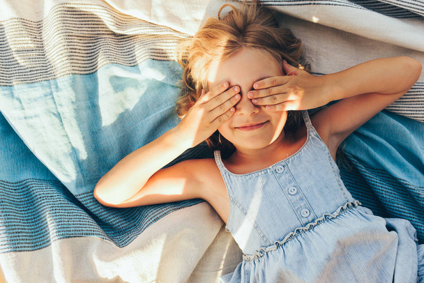 Beautiful blonde little girl lying on the blanket, cover her eyes from the sun with both hands, enjoying summer day. Adorable child having fun and plaiyng peekaboo outdoors in the park - Zdjęcie, obraz