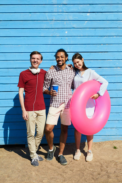 Pełna długość Portret trzech młodych ludzi stwarzających na plaży stoi przed niebieską ścianę i uśmiechnięty w aparacie, przestrzeń kopiowania - Zdjęcie, obraz