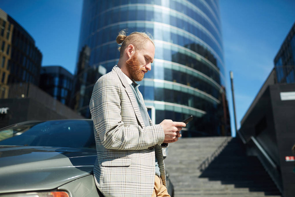 Серьезный вдумчивый хипстер молодой бизнесмен с хвостиком, опирающийся на автомобиль на улице и смс-кой по телефону
 - Фото, изображение