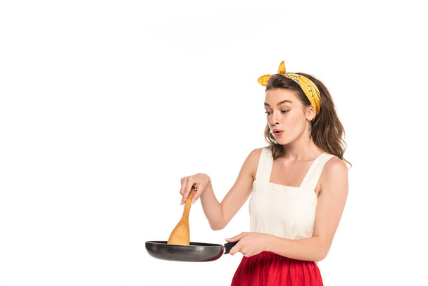 young shocked housewife in dress and apron looking at frying pan and wooden spatula isolated on white - Foto, afbeelding