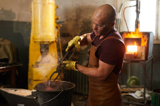 Serious bald blacksmith in apron and gloves using tongs while putting heated metal piece in bucket of water - Photo, Image
