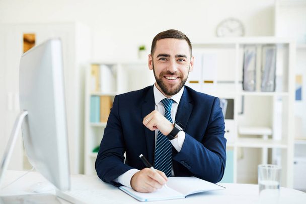 Portrait de heureux excité jeune PDG barbu en costume formel assis à la table avec ordinateur et écrire des objectifs dans le journal
 - Photo, image