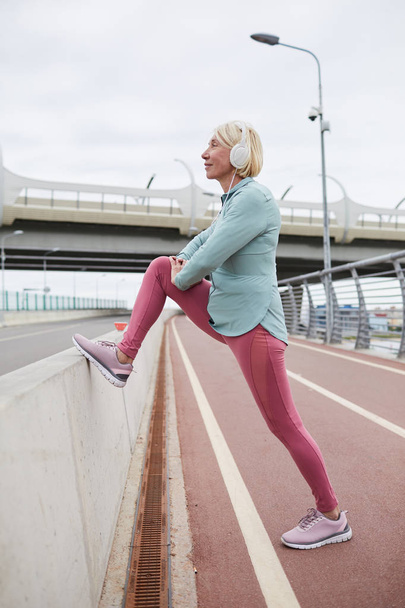 Активна зріла жінка в легінсах і вітровиках займається вправами для ніг розтягується, стоячи на гоночній доріжці на відкритому повітрі
 - Фото, зображення
