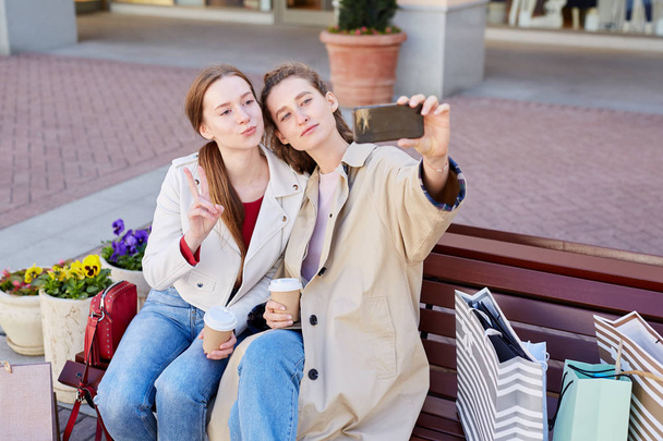Содержание стильные молодые женщины в повседневных нарядах сидя на скамейке с кофейными чашками и позируя для селфи, сделанного на смартфоне
 - Фото, изображение